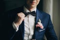 Stylish groom in blue suit with boutonniere with pink rose holding bow tie and posing near window in hotel room. Morning Royalty Free Stock Photo