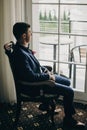 Stylish groom in blue suit, with bow tie and boutonniere with pink rose sitting in chair near window in hotel room. Morning Royalty Free Stock Photo