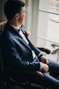 Stylish groom in blue suit, with bow tie and boutonniere with pink rose sitting in chair near window in hotel room. Morning Royalty Free Stock Photo