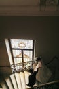 Stylish groom with gorgeous bride in amazing dress walking down stairs at window light in luxury room in hotel. rich wedding Royalty Free Stock Photo