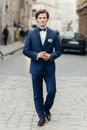 Stylish groom posing on background of old wall in city street. luxury handsome man in blue suit with flowers walking in street Royalty Free Stock Photo