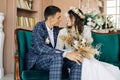 Stylish groom in a suit and a young bride in a wedding lace dress with a bouquet of flowers, sit together, hug, in the studio on Royalty Free Stock Photo