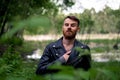 Stylish guy with a beard and mustache stands in a green forest against the background of grass and branches in a leather jacket wi Royalty Free Stock Photo