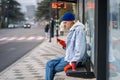 Stylish guy waiting bus sitting on bus stop bench in downtown with coffee browsing phone.