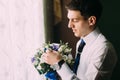 Stylish handsome dark haired groom holding a wedding bouquet standing near the window