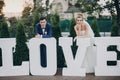 Stylish happy bride and groom posing at big love word in evening light at wedding reception outdoors. Gorgeous wedding couple of