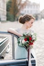 Stylish happy bride standing near the black retro car and holding wedding bouquet. Old city buildings on the backgroud Royalty Free Stock Photo