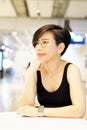 Stylish middle-age Asian woman sitting, thinking, smiling with hand on chin in modern cafe