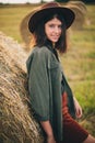 Stylish happy girl posing at hay bale in summer field in sunset. Portrait of young sensual woman in hat smiling at haystack, Royalty Free Stock Photo