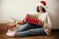 Stylish happy girl in santa hat and cozy sweater opening christmas gift box on background of presents in modern room. Young Royalty Free Stock Photo