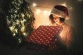 Stylish happy girl in santa hat and cozy sweater opening christmas gift box with magic light in dark room at modern christmas tree Royalty Free Stock Photo