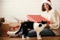 Stylish happy girl in santa hat and cozy sweater opening christmas gift box and playing with cats in decorated christmas room. Royalty Free Stock Photo