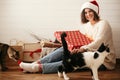 Stylish happy girl in santa hat and cozy sweater opening christmas gift box and playing with cats in decorated christmas room. Royalty Free Stock Photo