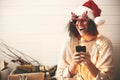 Stylish happy girl in santa hat and festive glasses with reindeer horns holding phone and smiling in christmas lights. Young Royalty Free Stock Photo