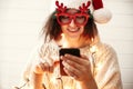 Stylish happy girl in santa hat and festive glasses with reindeer horns holding phone and smiling in christmas lights. Young Royalty Free Stock Photo