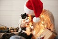 Stylish happy girl in santa hat kissing and playing with cute cat in festive christmas lights on background of modern presents. Royalty Free Stock Photo