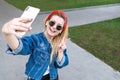 Stylish happy girl in sunglasses, a denim jacket and a scarf standing in the park, takes selfie on smartphone and smiling.Smiling Royalty Free Stock Photo