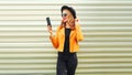 Stylish happy smiling woman taking selfie picture by smartphone wearing yellow jacket, black round hat on metal wall Royalty Free Stock Photo