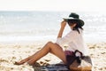 Stylish happy young woman relaxing on beach. Hipster girl in white shirt and hat sitting and tanning on beach near sea with waves Royalty Free Stock Photo