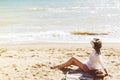 Stylish happy young woman relaxing on beach. Hipster girl in white shirt and hat sitting and tanning on beach near sea with waves Royalty Free Stock Photo