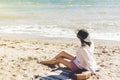 Stylish happy young woman relaxing on beach. Hipster girl in white shirt and hat sitting and tanning on beach near sea with waves Royalty Free Stock Photo