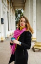 Happy young woman is wearing a pink scarf, She is holding a coffee to go. Portrait of smiling girl in autumn city. Royalty Free Stock Photo