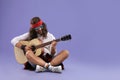 Stylish hippie man with guitar on violet background, space for text