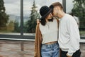 Stylish hipster couple hugging on wooden porch, relaxing in modern cabin in mountains. Happy young family in modern outfits