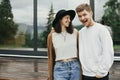 Stylish hipster couple smiling on wooden porch, relaxing in modern cabin in mountains. Happy young family in modern outfits