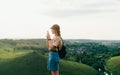 Stylish hipster girl with a backpack on an expedition in mountains stands on edge of a cliff and uses a smartphone. Attractive