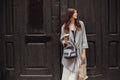 Stylish hipster girl with beautiful hair and smile holding coffee cup and paper bag, standing at old wooden door in city street . Royalty Free Stock Photo