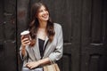 Stylish hipster girl with beautiful hair and smile holding coffee cup and paper bag, standing at old wooden door in city street . Royalty Free Stock Photo