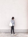 Stylish hipster girl in denim outfit and tote bag posing in city street on background of wall. Phone photo. Fashionable woman with