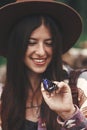 Stylish Hipster Girl In Hat Holding Purple Emperor Butterfly On Hand In Summer Mountains. Young Woman Traveler With Butterfly,