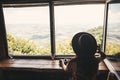 Stylish hipster girl in hat looking on top of mountains through big window in old cabin. Young woman traveler relaxing in summer Royalty Free Stock Photo