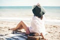 Stylish hipster girl in hat sitting on beach with straw bag and tanning near sea waves. Summer vacation. Happy boho woman relaxing Royalty Free Stock Photo