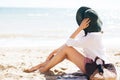 Stylish hipster girl in hat sitting on beach with straw bag and tanning near sea waves. Summer vacation. Happy boho woman relaxing Royalty Free Stock Photo