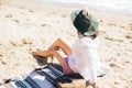 Stylish hipster girl in hat sitting on beach with straw bag and tanning. Summer vacation. Happy young boho woman relaxing and Royalty Free Stock Photo