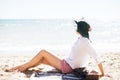 Stylish hipster girl in hat sitting on beach and tanning near sea waves. Summer vacation. Happy boho woman relaxing and enjoying Royalty Free Stock Photo