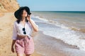 Stylish hipster girl in hat walking on beach and smiling. Summer vacation. Happy young boho woman relaxing and enjoying sunny warm Royalty Free Stock Photo