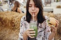 Stylish hipster girl holding delicious vegan burger and drinking smoothie in glass jar at street food festival. Happy boho woman