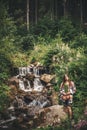 Stylish Hipster Girl Holding Fern Leaf And Relaxing In Forest In Mountains. Young Woman Traveler Exploring Waterfall Woods. Eco