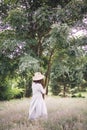 Stylish hipster girl in linen dress and hat relaxing in lavender field near tree. Happy bohemian woman enjoying summer vacation in