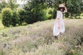 Stylish hipster girl in linen dress and hat relaxing in lavender field near tree. Happy bohemian woman enjoying summer vacation in