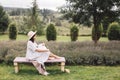 Stylish hipster girl in linen dress and hat sitting on bench at lavender field and relaxing in the morning. Happy bohemian woman