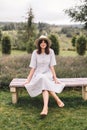 Stylish hipster girl in linen dress and hat sitting on bench at lavender field and relaxing in the morning. Happy bohemian woman