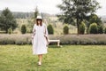 Stylish hipster girl in linen dress and hat walking at lavender field and relaxing in mountains. Bohemian woman smiling and