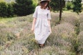 Stylish hipster girl in linen dress and hat walking in lavender field and smiling. Happy bohemian woman relaxing and enjoying