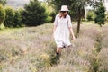 Stylish hipster girl in linen dress and hat walking in lavender field and smiling. Happy bohemian woman relaxing and enjoying