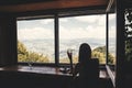 Stylish hipster girl looking on top of mountains through big window in old cabin. Young woman relaxing with drink in summer Royalty Free Stock Photo